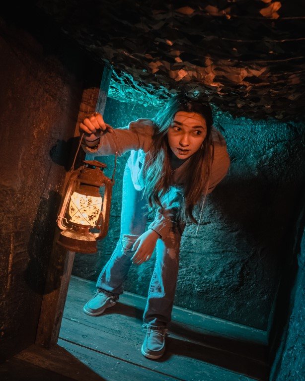 Young woman holding a lantern leans through a narrow, blue-lit tunnel passage