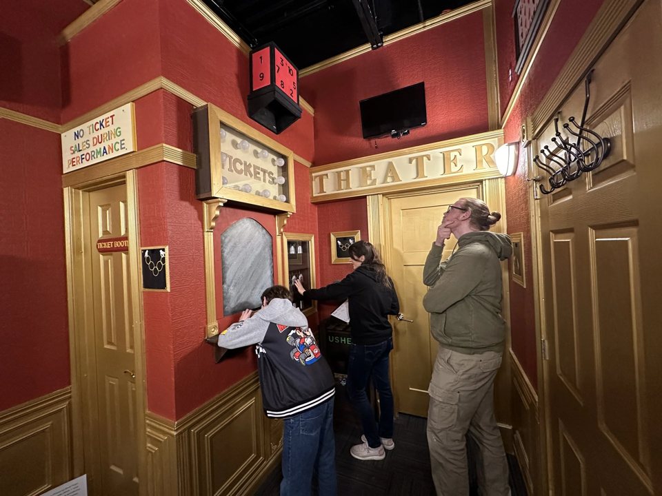 Three players work on puzzles in a red and gold theater lobby set next to a ticket booth
