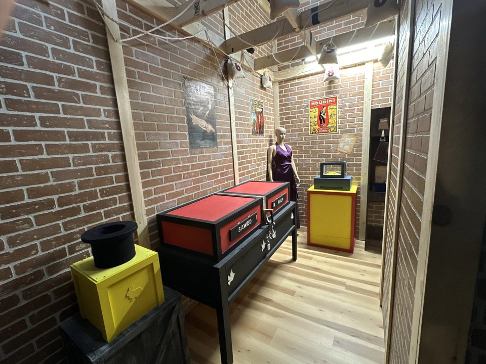Brick-walled magic room with a sawing-in-half box, a top hat and a Houdini poster