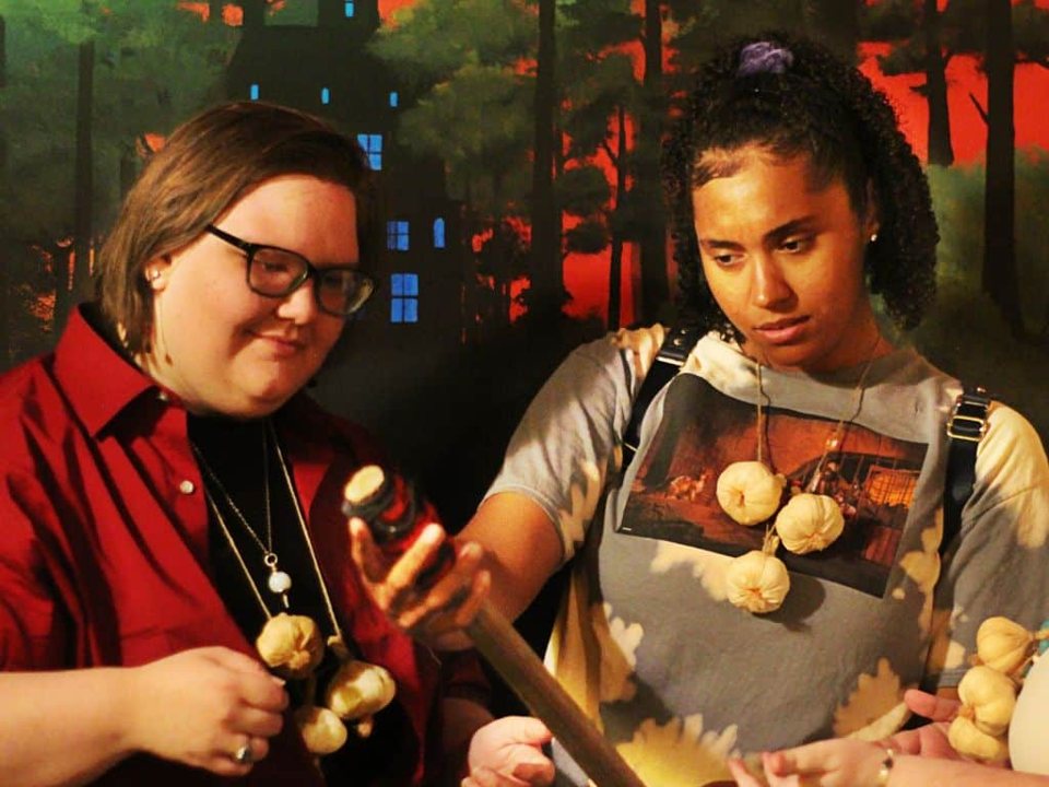 Two players wearing garlic necklaces examine a bottle in front of a haunted forest mural