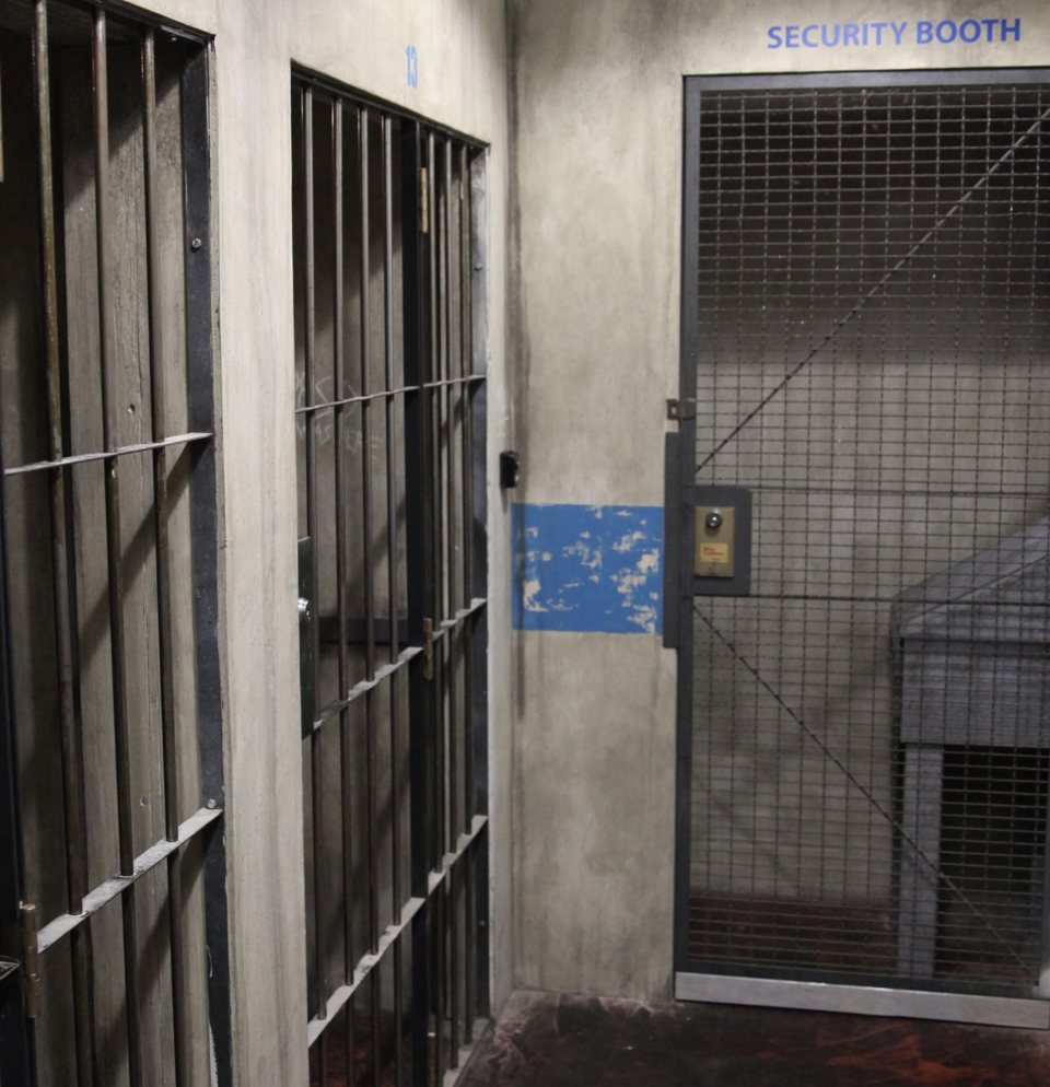 Rácsos börtöncellák és drótrácsos biztonsági fülke egy kopott betonfolyosón