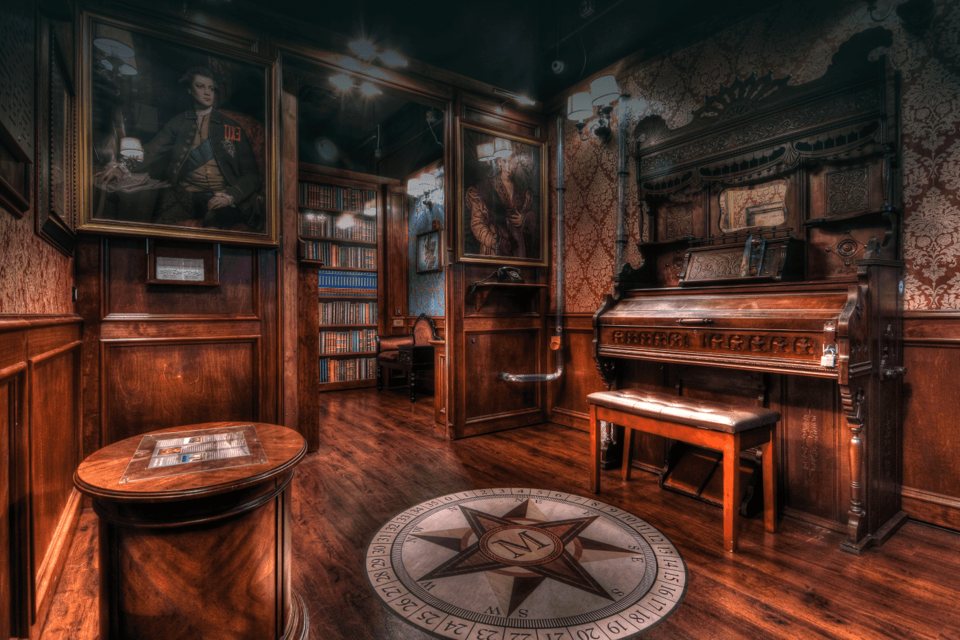 Wood-paneled Victorian parlor set with an antique pump organ, oil portraits, bookshelves and a compass rose on the floor