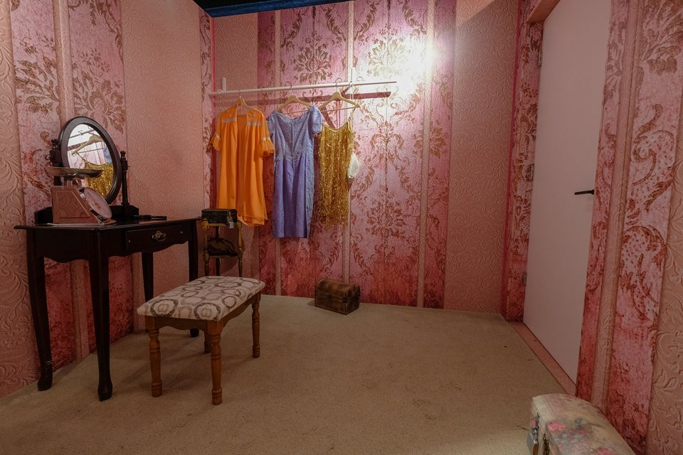 Pink dressing room with a vanity, an upholstered bench and vintage dresses on a clothes rail