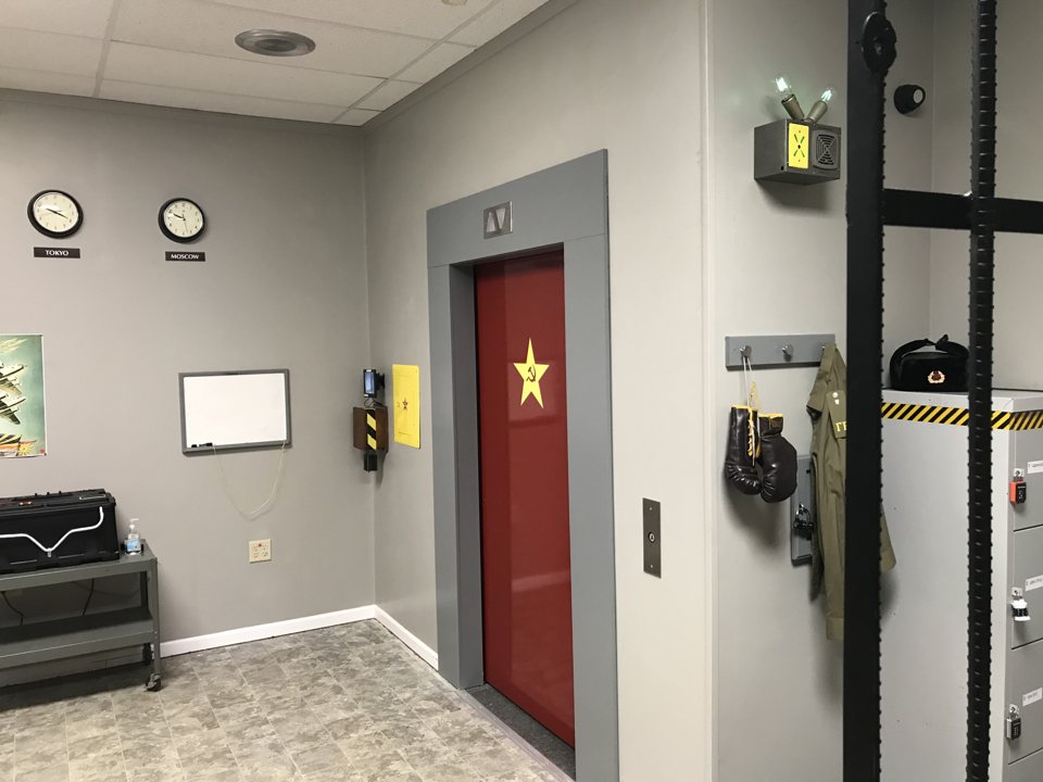 Cold War spy room with Tokyo and Moscow clocks, a red elevator door with a Soviet star and a uniform on a hook