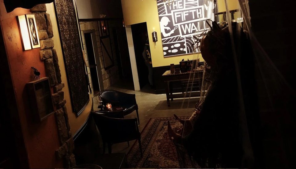 Dim lobby with armchairs, a Persian rug, a stone wall and a large Fifth Wall mural, with a costumed figure behind a veil in the foreground