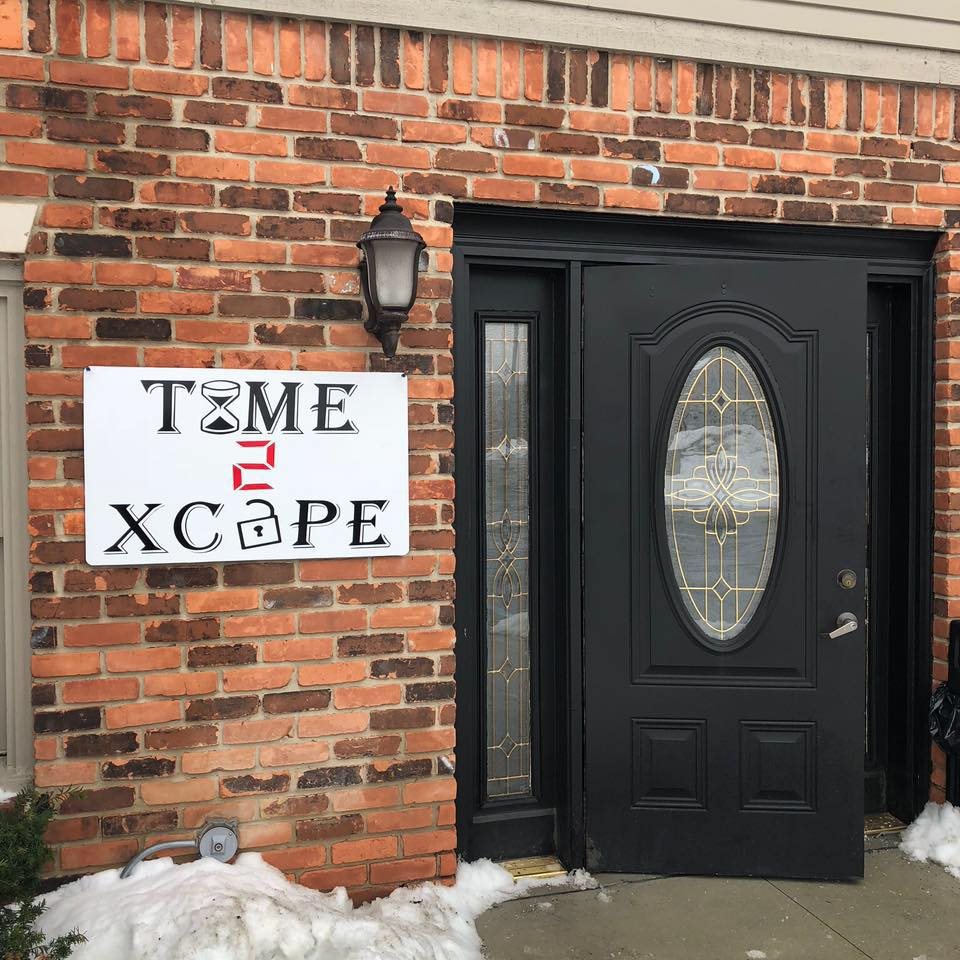 Black entrance door in a brick wall with the Time 2 Xcape sign and a wall lamp beside it and snow piled in front