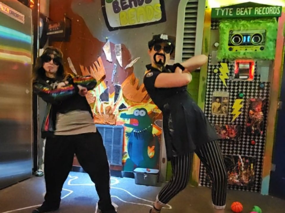 Two costumed players in sunglasses pose in the colorful retro-style room in front of the Tyte Beat Records set