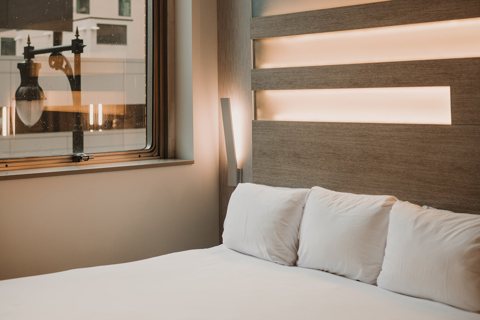 A guest room with a lit wooden headboard and a street-facing window