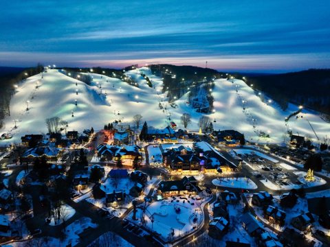 Lit ski slopes and the snowy resort village in an evening aerial view