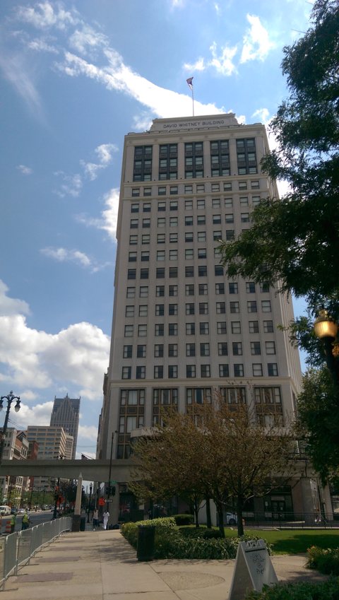 The tall, pale facade of the David Whitney Building under a blue sky, seen from Woodward Avenue