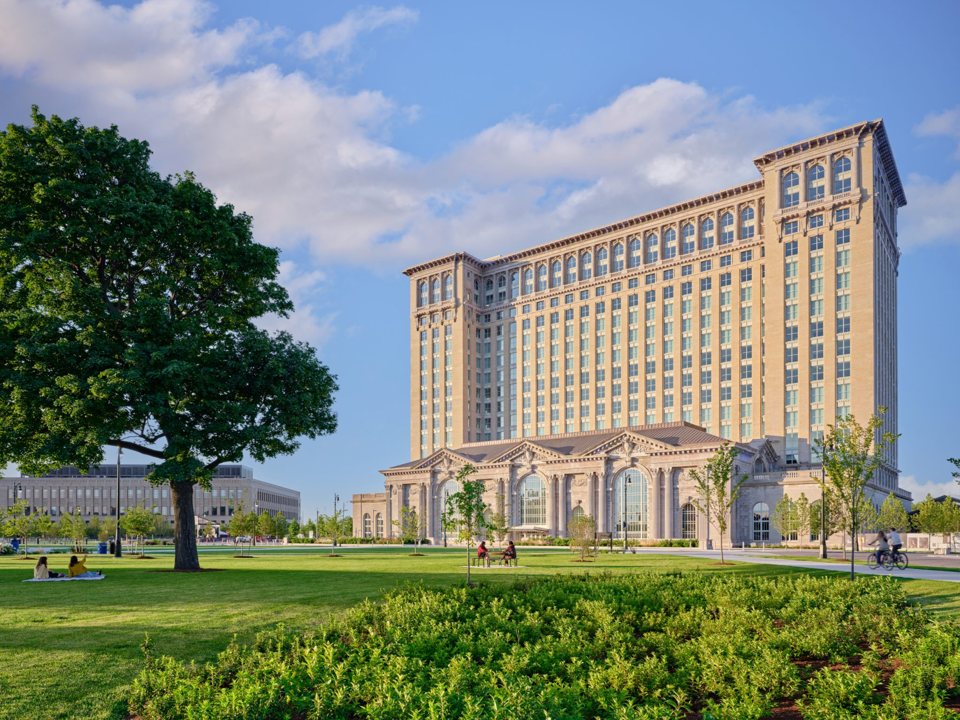 The restored Michigan Central Station tower and depot building in sunshine, with a leafy park in front