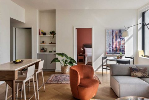 A sunny apartment living room with a kitchen counter, an orange armchair and a door to the bedroom