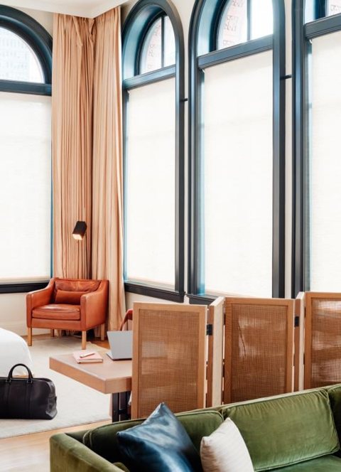 A guest room with tall arched windows, a leather armchair, a green velvet sofa and a woven screen