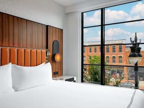 A guest room with a king bed, a wooden headboard and a large window overlooking nearby brick buildings