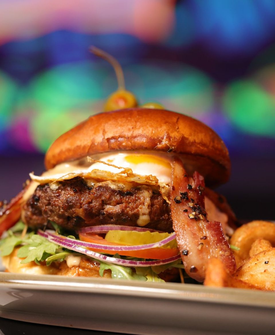 A burger with a fried egg, bacon, tomato and red onion in front of colorful lights