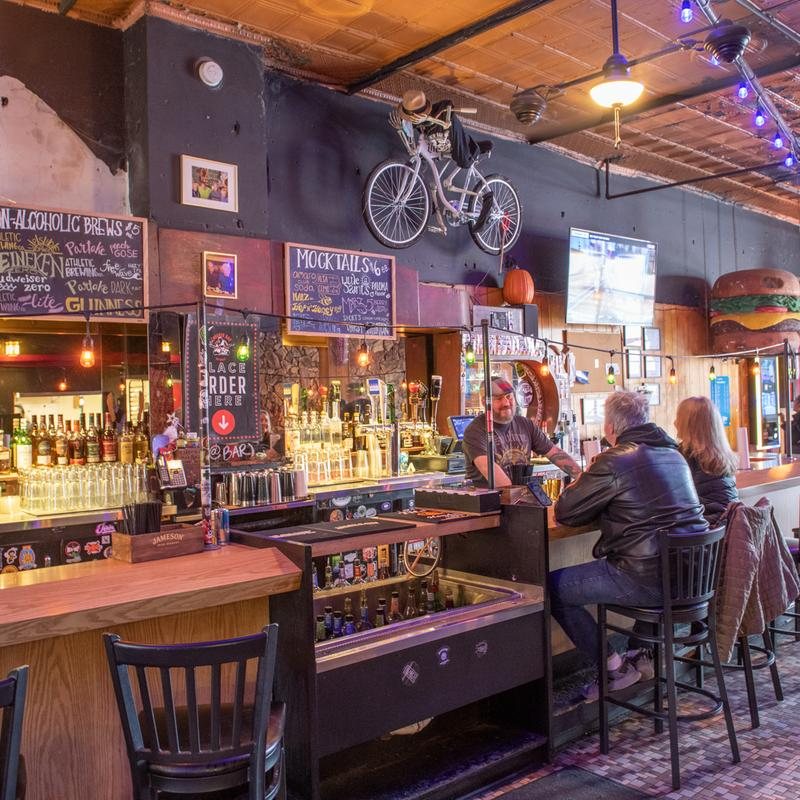 Back bar shelves with chalkboard drink menus, a bicycle above them and guests sitting at the bar
