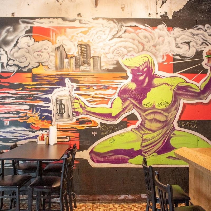 A mural echoing the Spirit of Detroit statue holding a beer mug in front of the Detroit skyline, with tables below