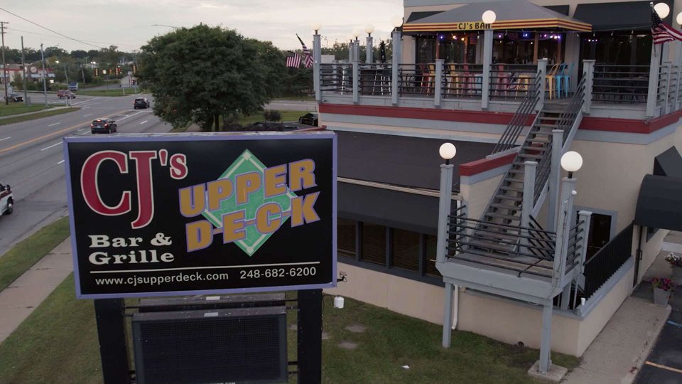 The roadside CJ's Upper Deck sign with the building's upstairs deck and staircase behind it