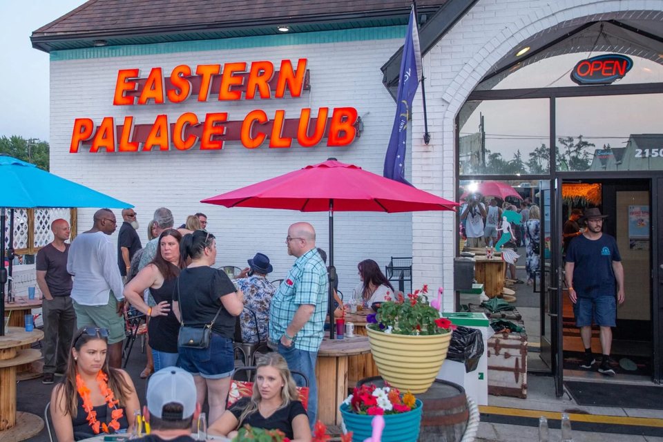 A white building with a red Eastern Palace Club sign and a patio of chatting guests under umbrellas