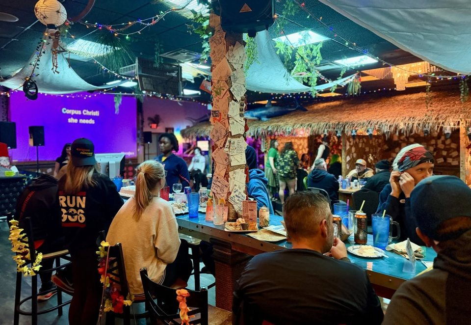 Guests at long tables beside a thatched bar with string lights and a purple-lit screen in the background