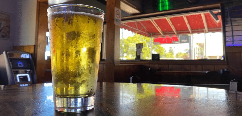 A glass of light beer on a glossy bar table, with a window and a red awning in the background