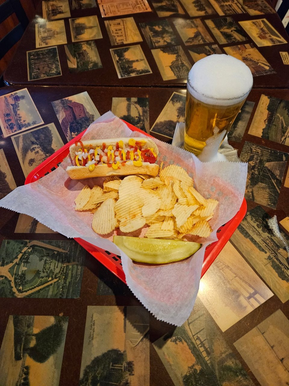 A hot dog with mustard and ketchup, ridged chips and a pickle in a red basket, next to a glass of beer