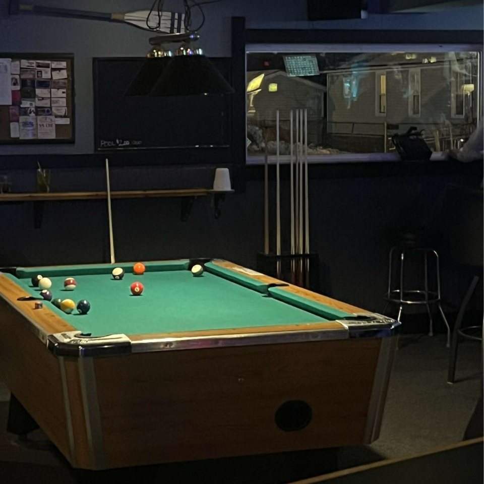 A pool table in the corner of the bar with a cue rack on the wall