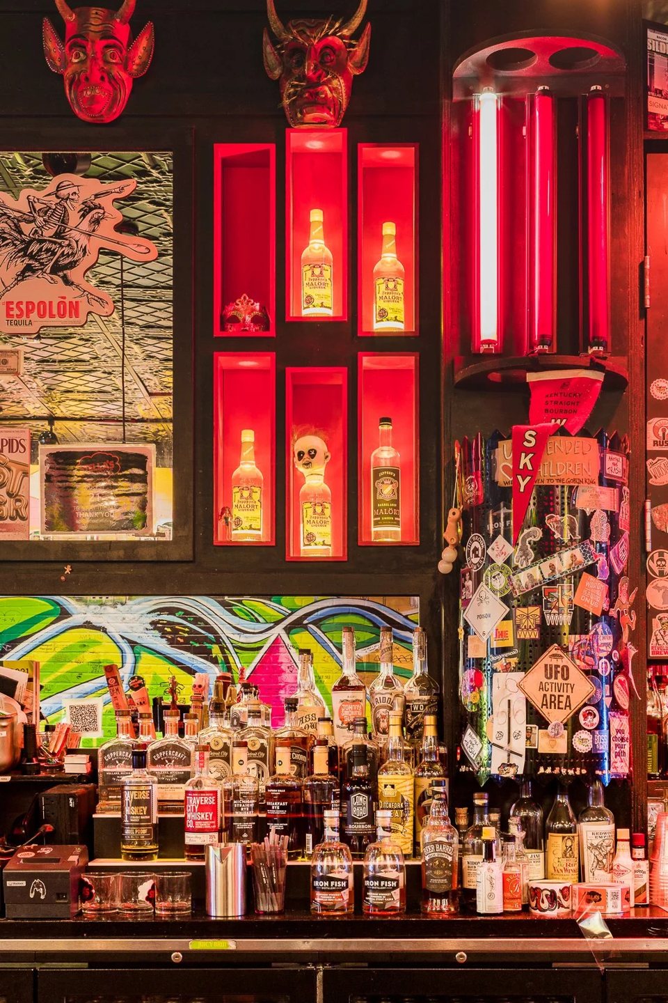 Liquor shelves glowing in red light, masks and sticker-covered walls behind the bar