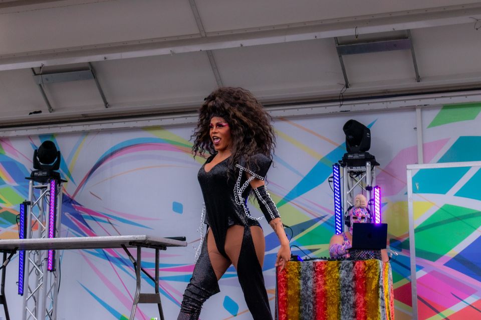 Drag-előadó fekete dresszben és csizmában egy színes hátterű színpadon