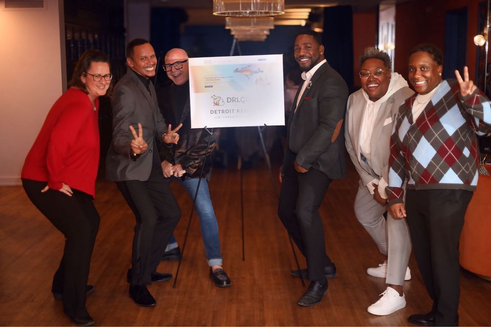 Cheerful chamber members pose beside an event sign in a chandelier-lit hall
