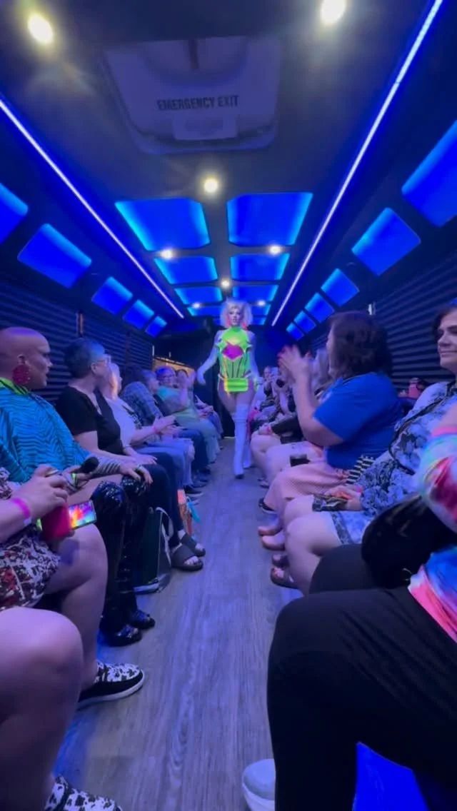 Blue-lit party bus interior with a drag performer walking the aisle between guests