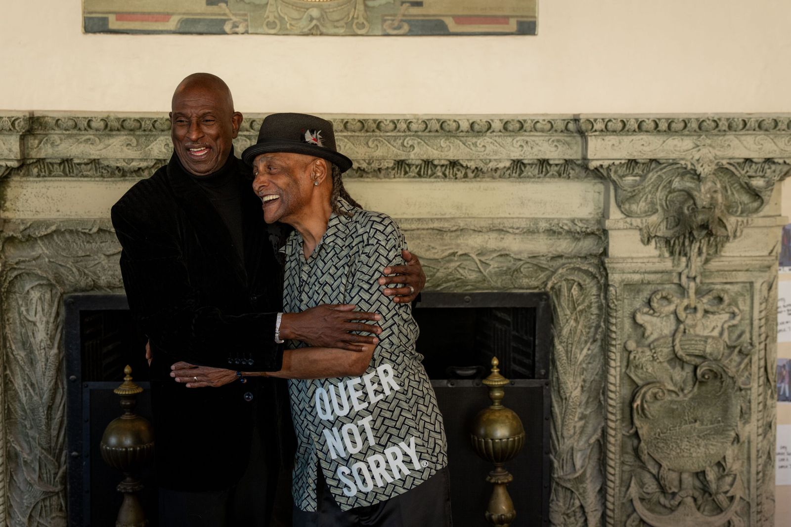 Two older people embrace, one carrying a 'Queer Not Sorry' tote bag