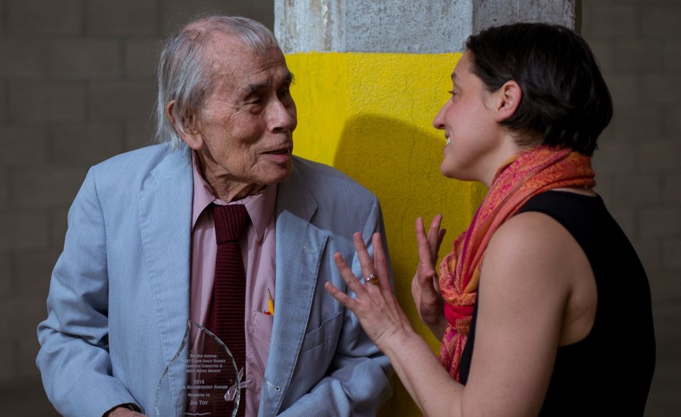 An older man holding an award chats with a younger woman