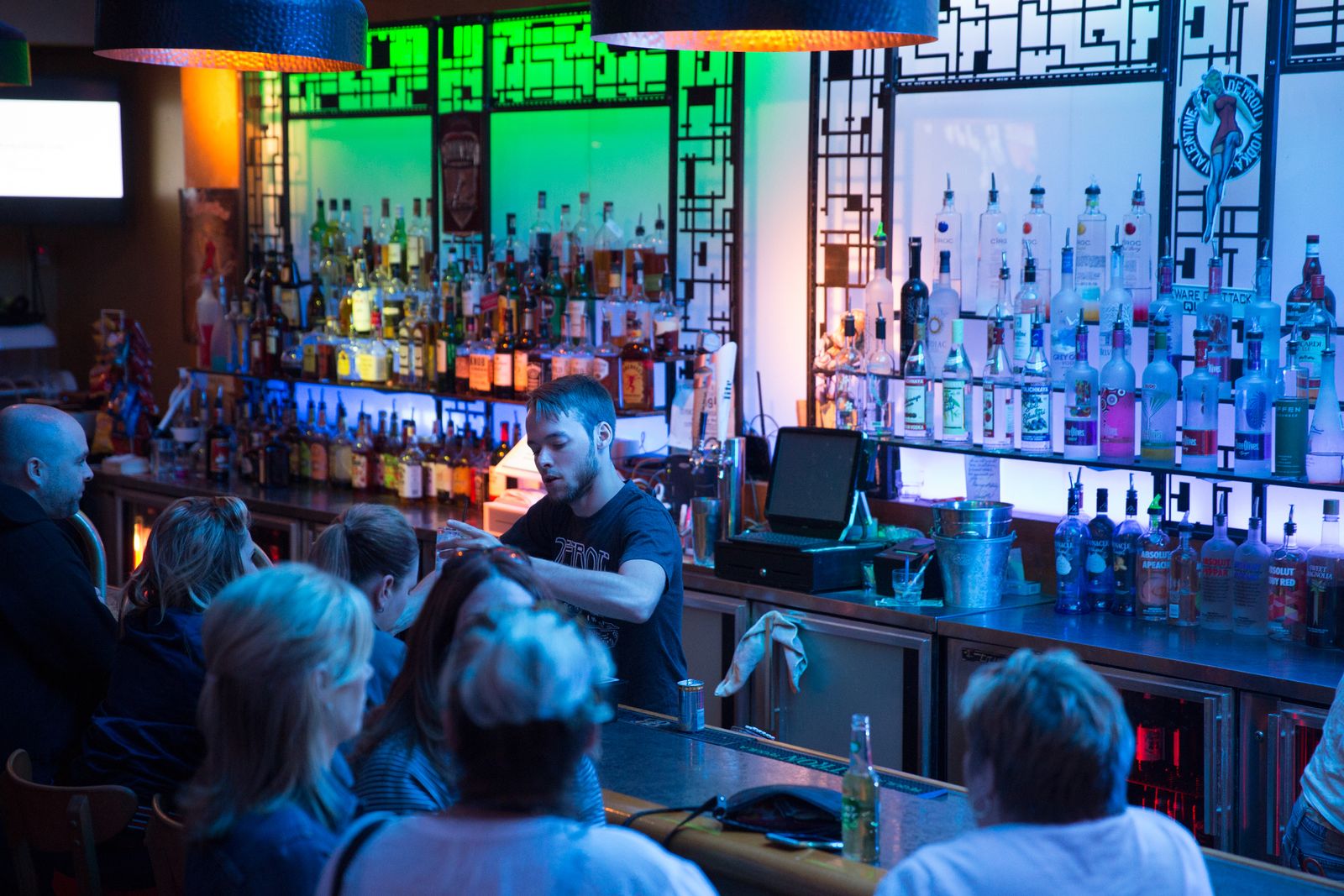 Bartender mixing drinks behind the colorfully lit back bar as guests look on