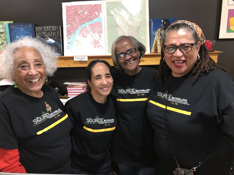 Four women in Source Booksellers shirts in front of the shop's shelves and wall maps