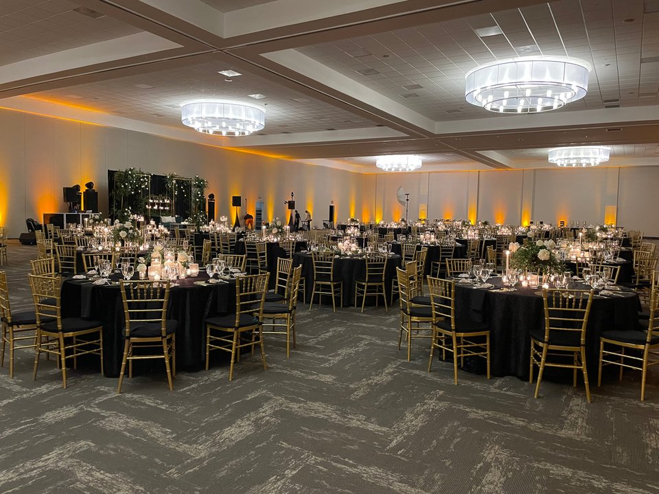 A ballroom set for dinner with black linens, gold chairs and round chandeliers