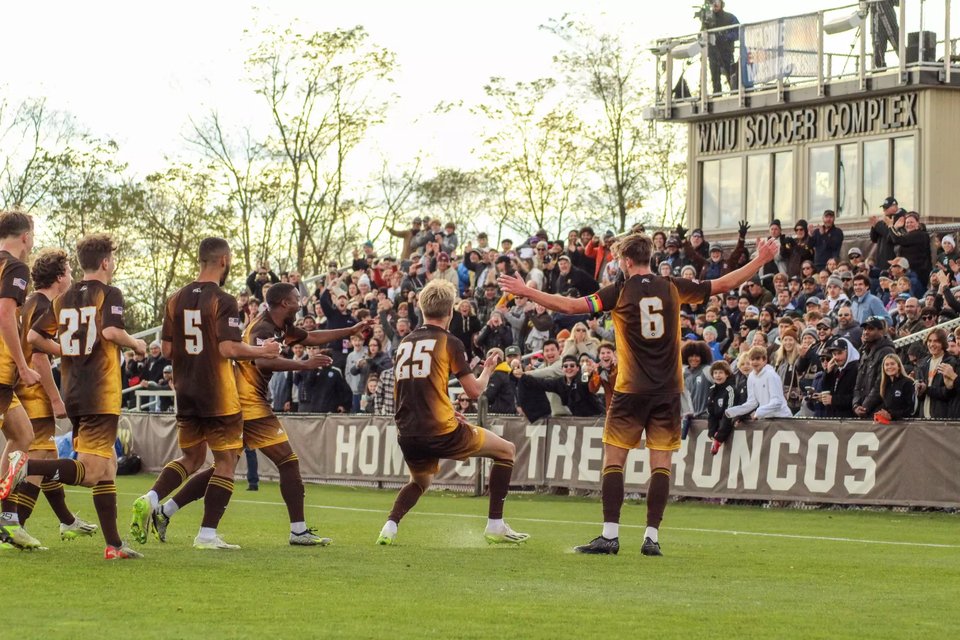 Bronco men's players celebrating a goal in front of the stands, press box above
