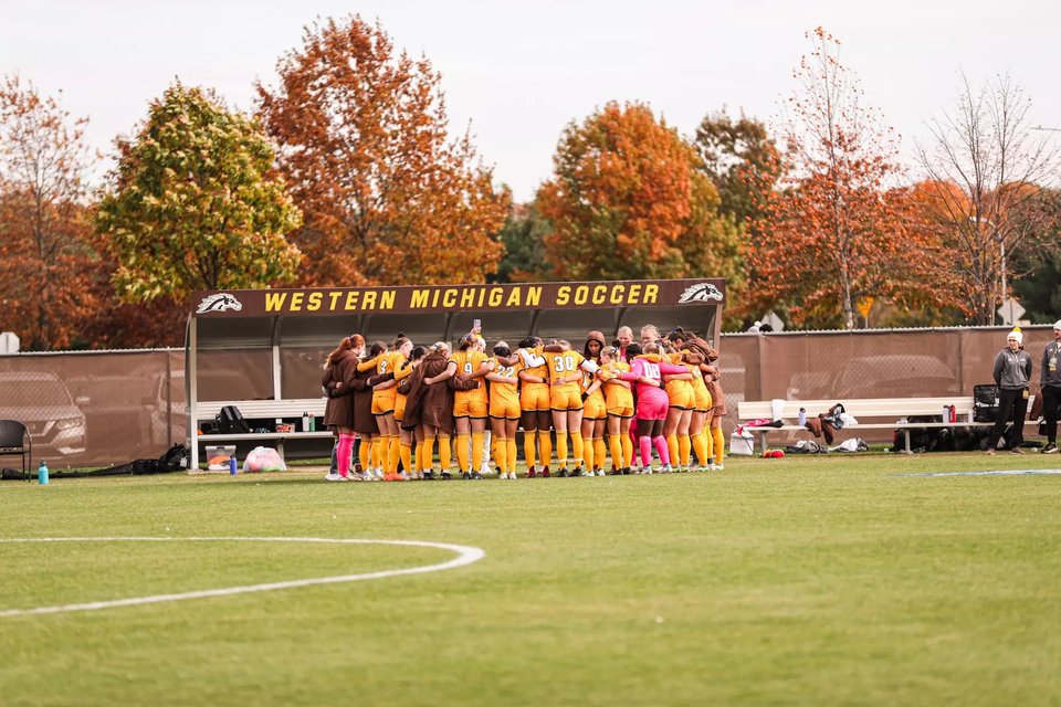The women's team huddled by the Western Michigan Soccer bench shelter, fall trees behind
