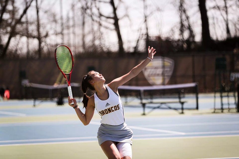 A Bronco women's player serving on an outdoor court, windscreens and benches behind