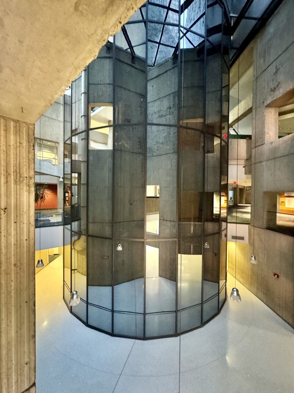 A glass-walled cylindrical elevator shaft among raw-concrete walls (2024)