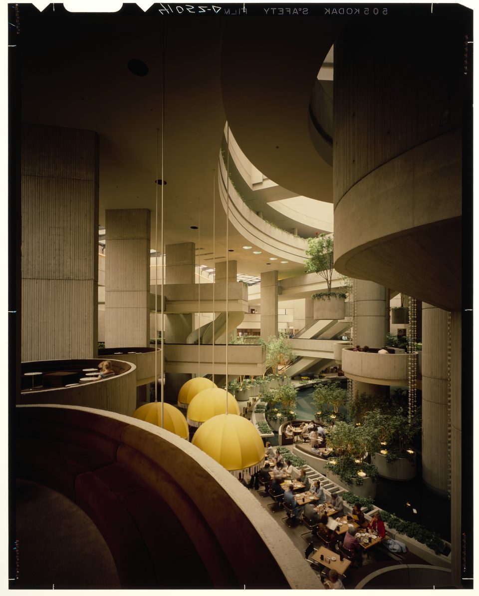 A restaurant under yellow umbrella lamps in the central atrium, curving concrete galleries above, 1977: a photo by Budapest-born Balthazar Korab