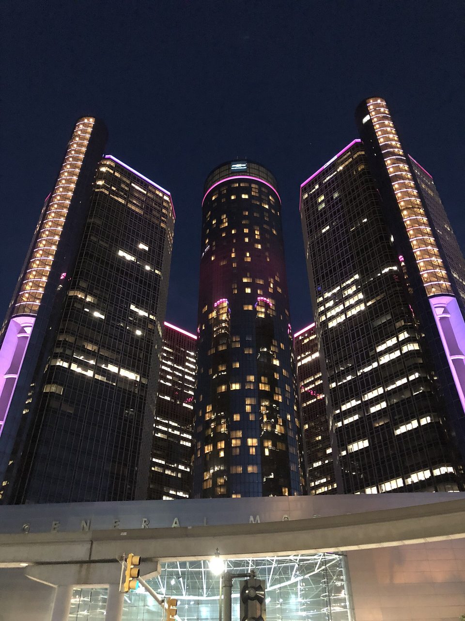 The towers with their purple light strips at night from the street, October 2018, above the entrance lettered “General Motors”