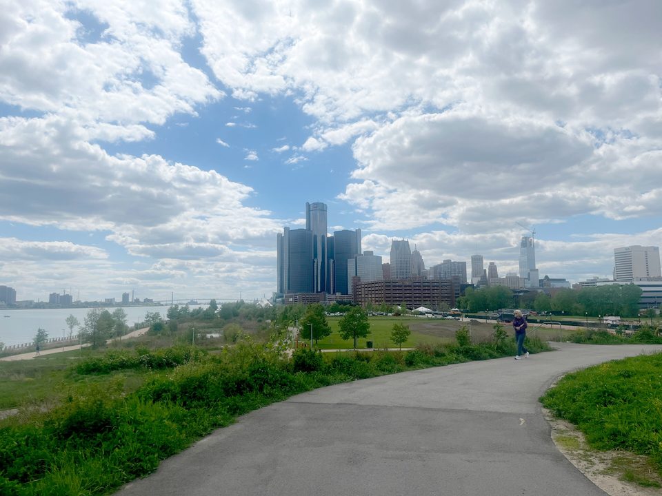 The east stretch of the RiverWalk in May 2024: open green riverfront with the Renaissance Center behind