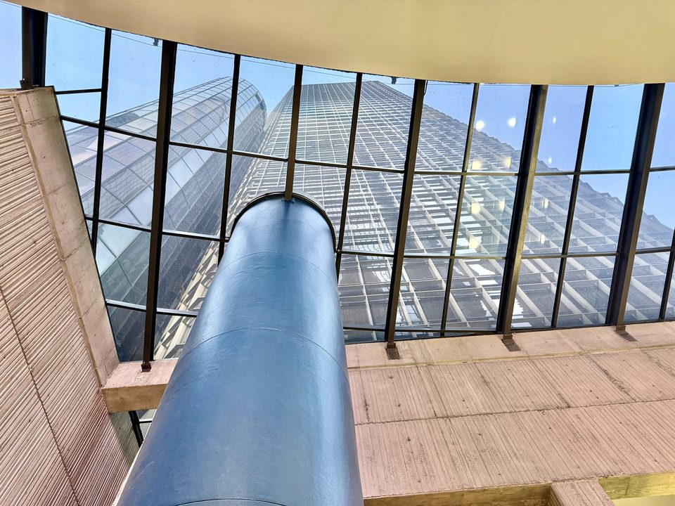 A skylight above the lobby, a blue cylinder rising through it and a tower’s glass wall beyond (2024)