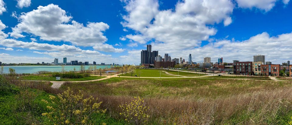 A panorama from the east stretch of the RiverWalk in April 2026: grassy riverbank, with downtown and the Renaissance Center beyond