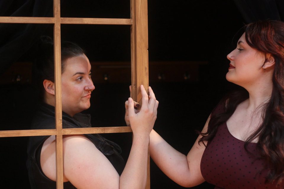 Two actresses face each other across a wooden window frame on a dark stage