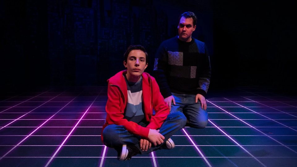A boy in a red hoodie sits cross-legged on a glowing grid-patterned stage, with a kneeling man behind him