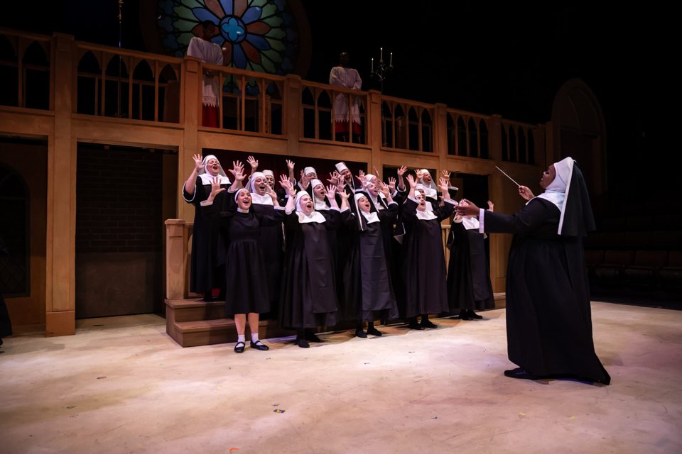A choir of nuns sings with raised hands in front of a conducting nun, on a church set with a rose window