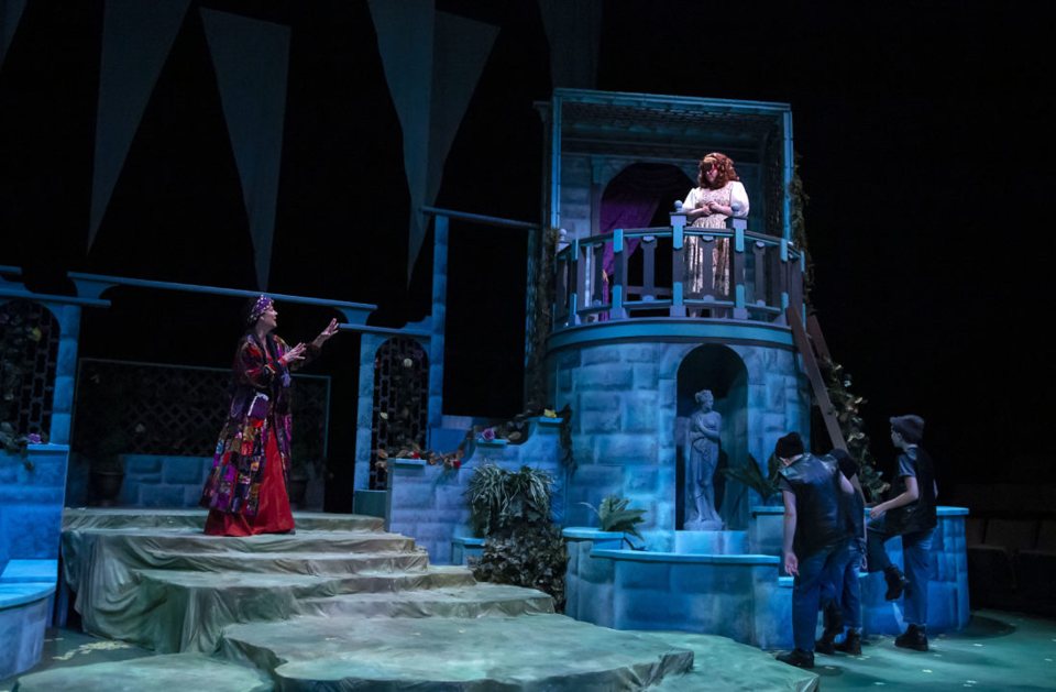 A woman in a patchwork coat calls up to a girl on the balcony of a blue stone tower set while boys watch from the right