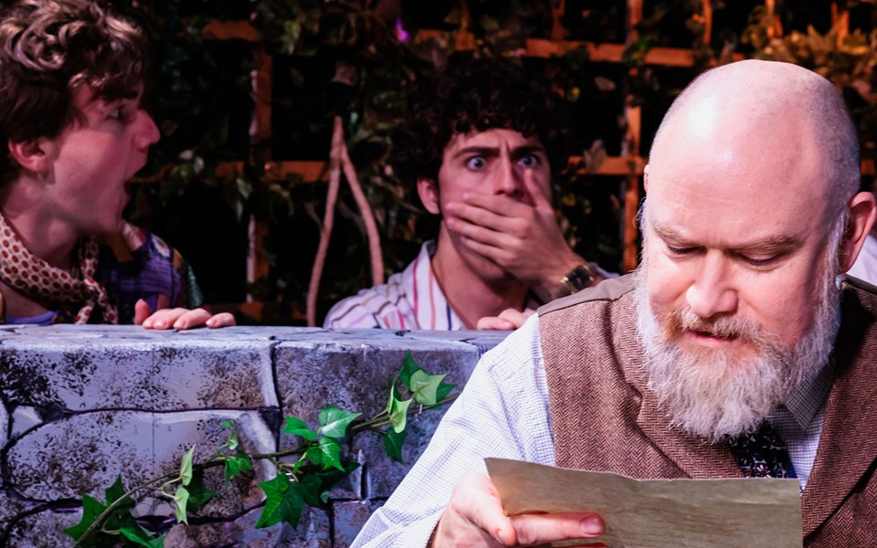 A bearded actor in a waistcoat reads a letter while three men peek out in shock from behind an ivy-covered stone wall